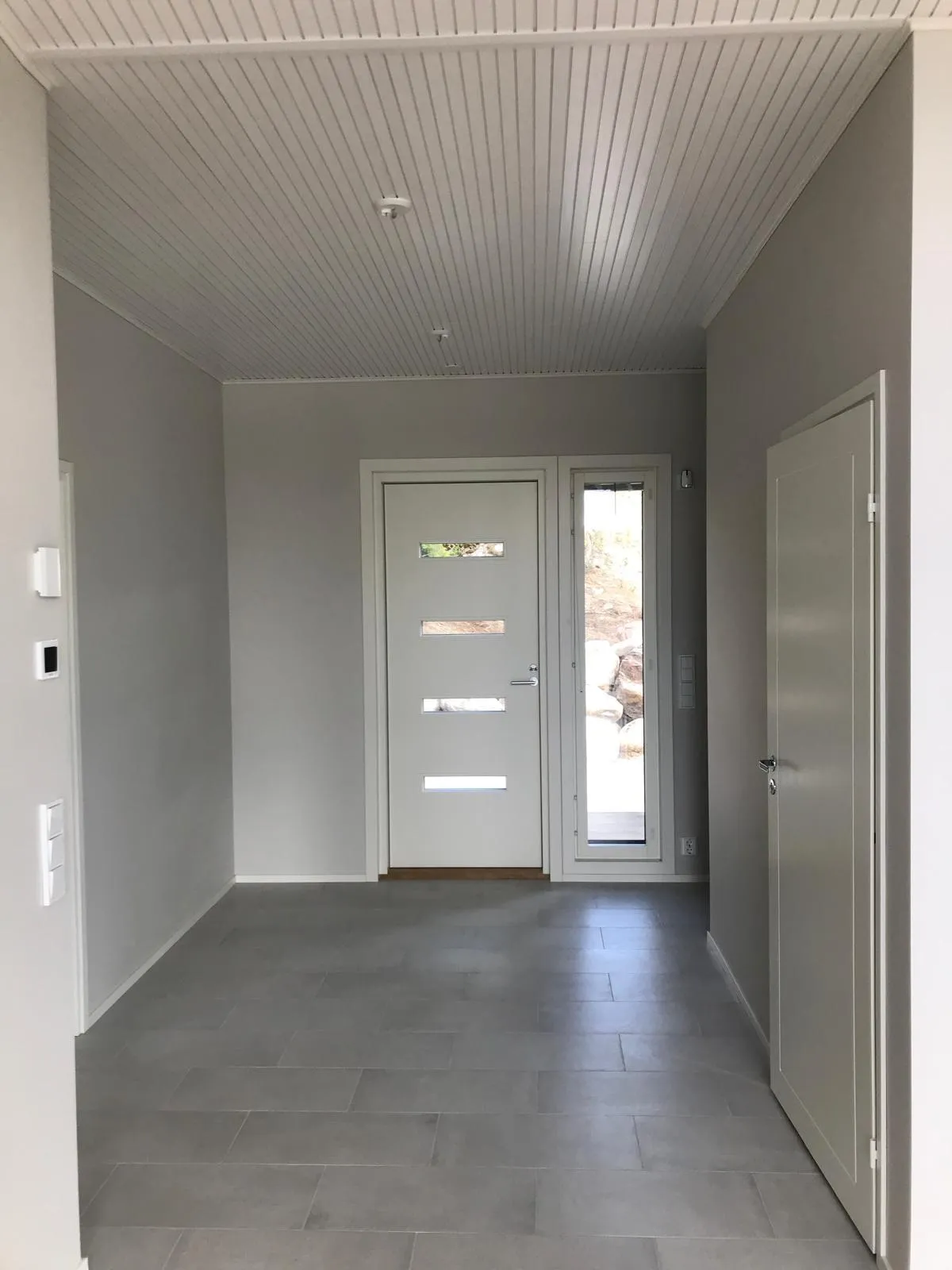 Sisäänkäynti harmaalla laattalattialla, valkoisella katolla ja valkoisilla ovilla.