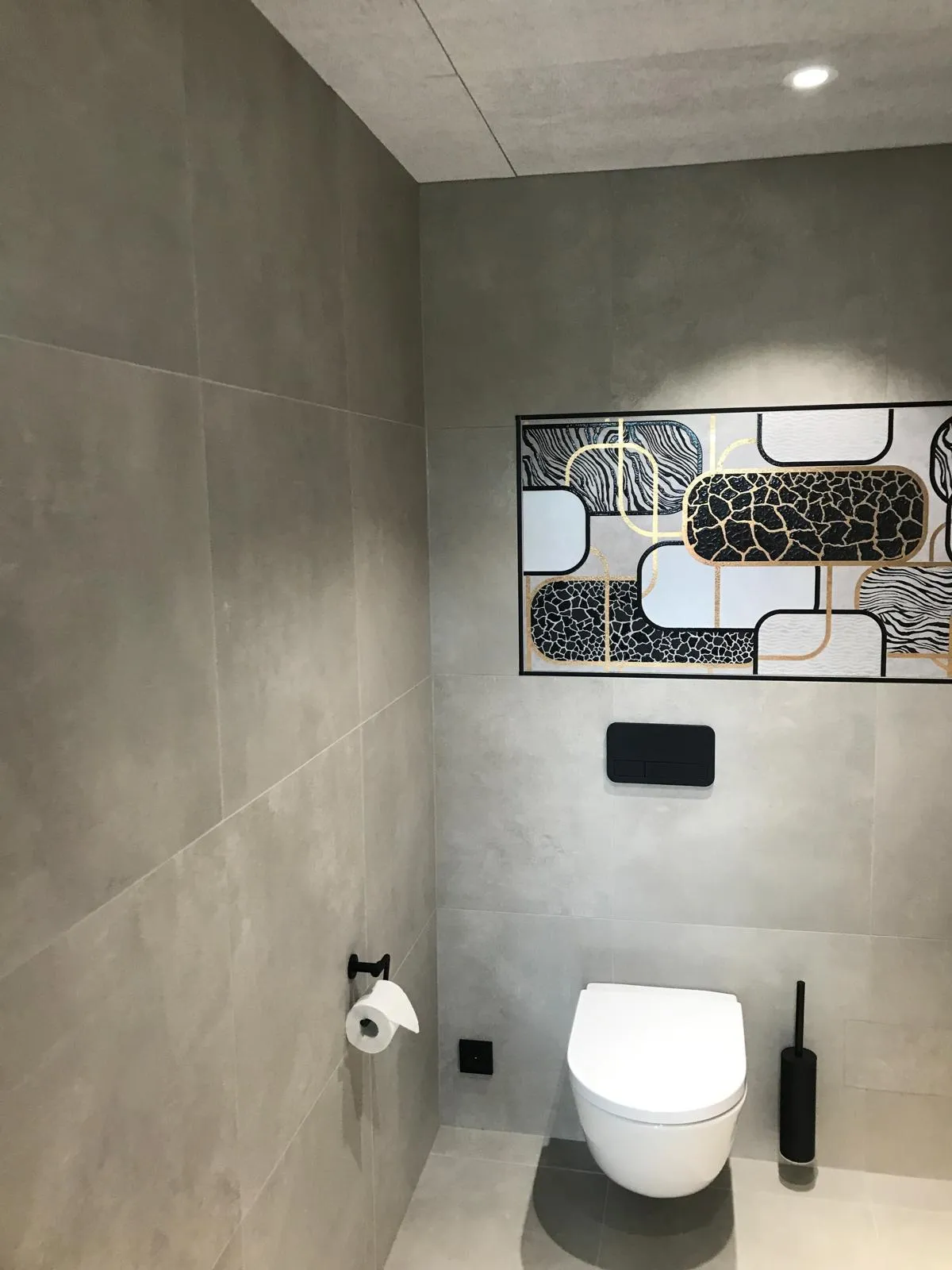 Harmaa kylpyhuone, jossa on seinällä taulu, valkoinen WC-istuin ja musta WC-harja.