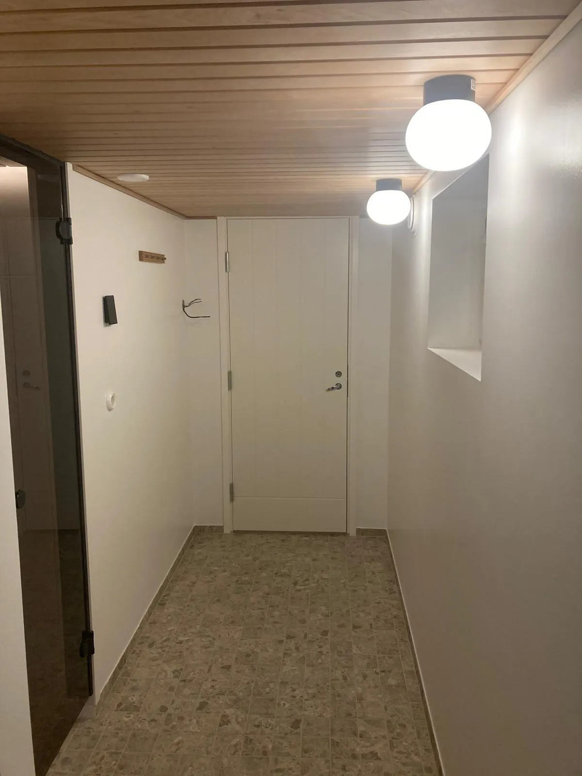 Valkoinen käytävä, jossa on puukatto, valkoiset seinät, kaksi pyöreää valaisinta ja ruskea laattalattia.