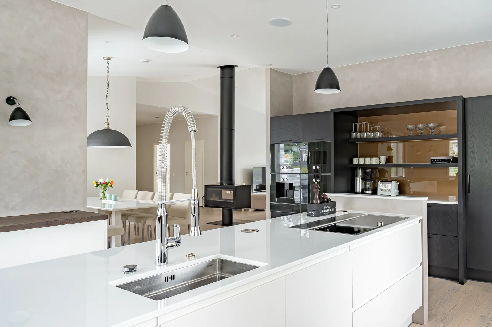 Moderni keittiö, jossa on valkoinen saareke, teräksinen pesuallas, mustat kaapit ja takka. Kattovalaisimet valaisevat tilaa.