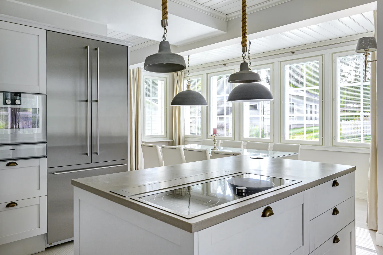 Vaaleassa keittiössä on ruostumattomasta teräksestä valmistettu jääkaappi, valkoinen keittiösaareke, jossa on harmaa työtaso ja sisäänrakennettu liesi, sekä roikkuvat valaisimet.