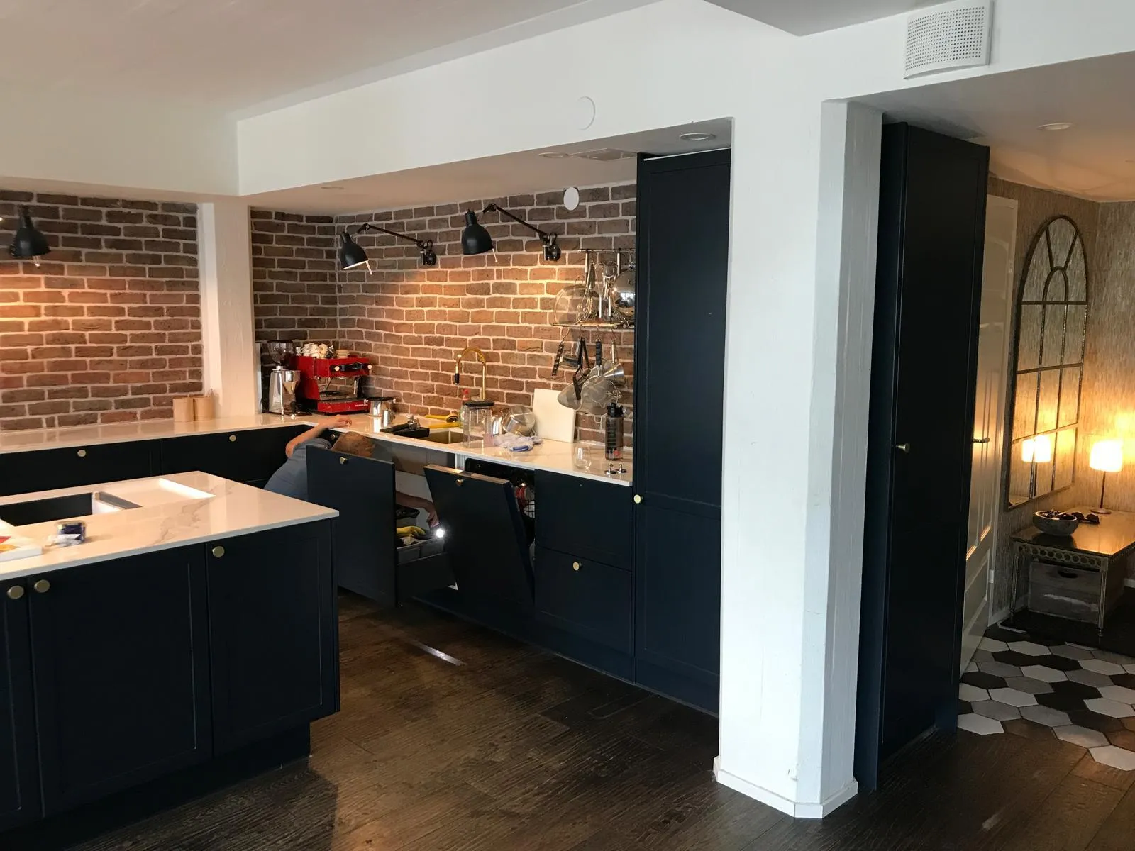 Keittiö, jossa on tummansiniset kaapit, tiiliseinä, valkoinen työtaso ja henkilö kumartuneena avoimen kaapin alle.