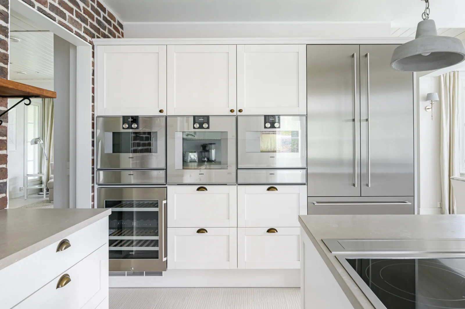 Keittiön sisustus valkoisilla kaapeilla, ruostumattomasta teräksestä valmistetuilla laitteilla ja harmaalla betonivalaisimella.