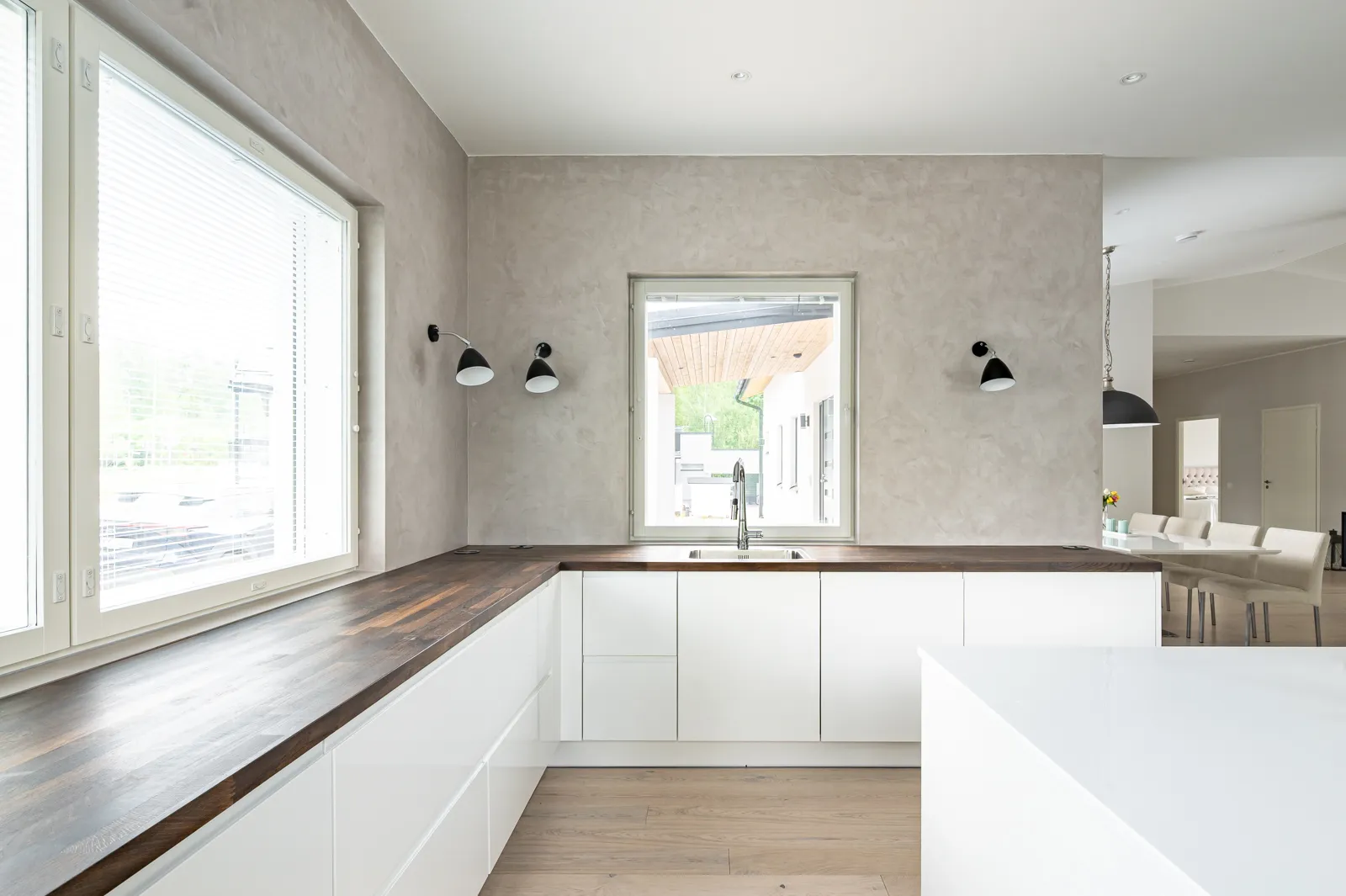 Moderni keittiö, jossa on valkoiset kaapit, tumma puutaso, kaksi ikkunaa ja mustat seinävalaisimet. Ruokailutila näkyy taustalla.