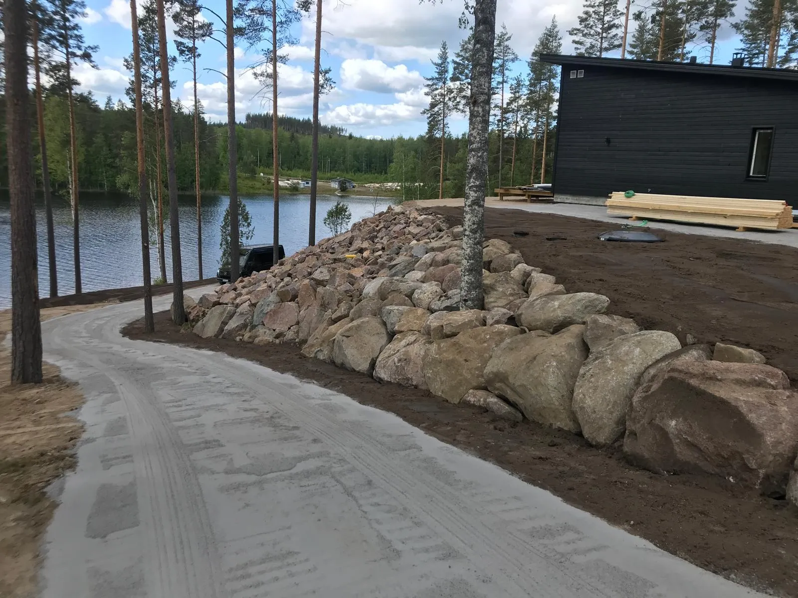 Maisema, jossa on kivimuuri, hiekkatie, järvi ja tumma puutalo aurinkoisena päivänä.