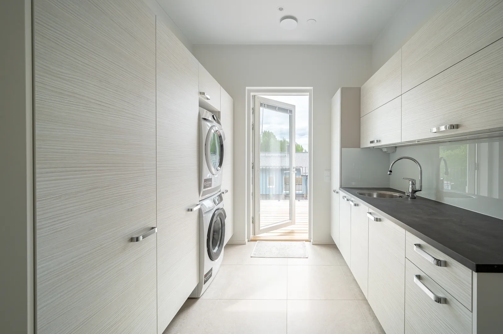 Vaaleassa kodinhoitohuoneessa on valkoiset kaapit, pesukone ja kuivausrumpu, tumma työtaso ja avoin ovi ulos.