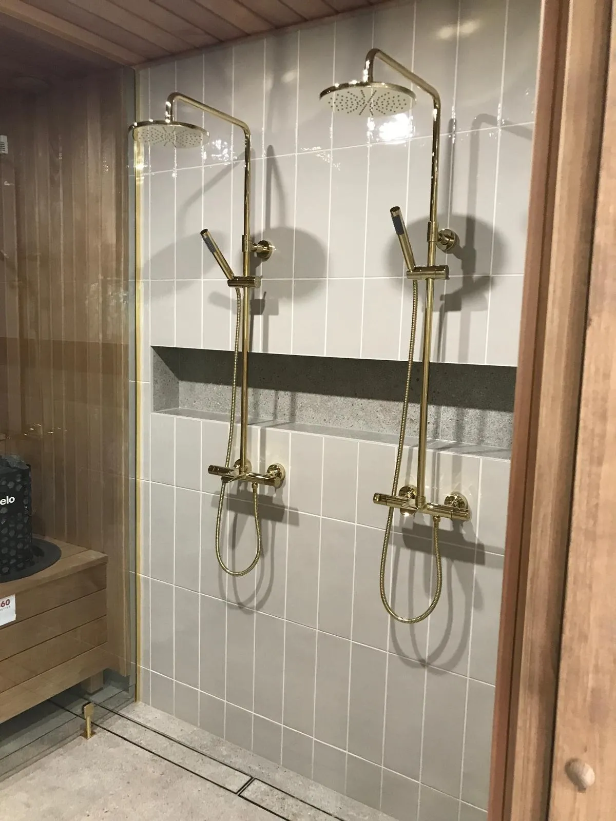 Kaksi kultaista suihkua harmaalla laattaseinällä, joissa on yläsuihku ja käsisuihku, sekä upotettu hylly.