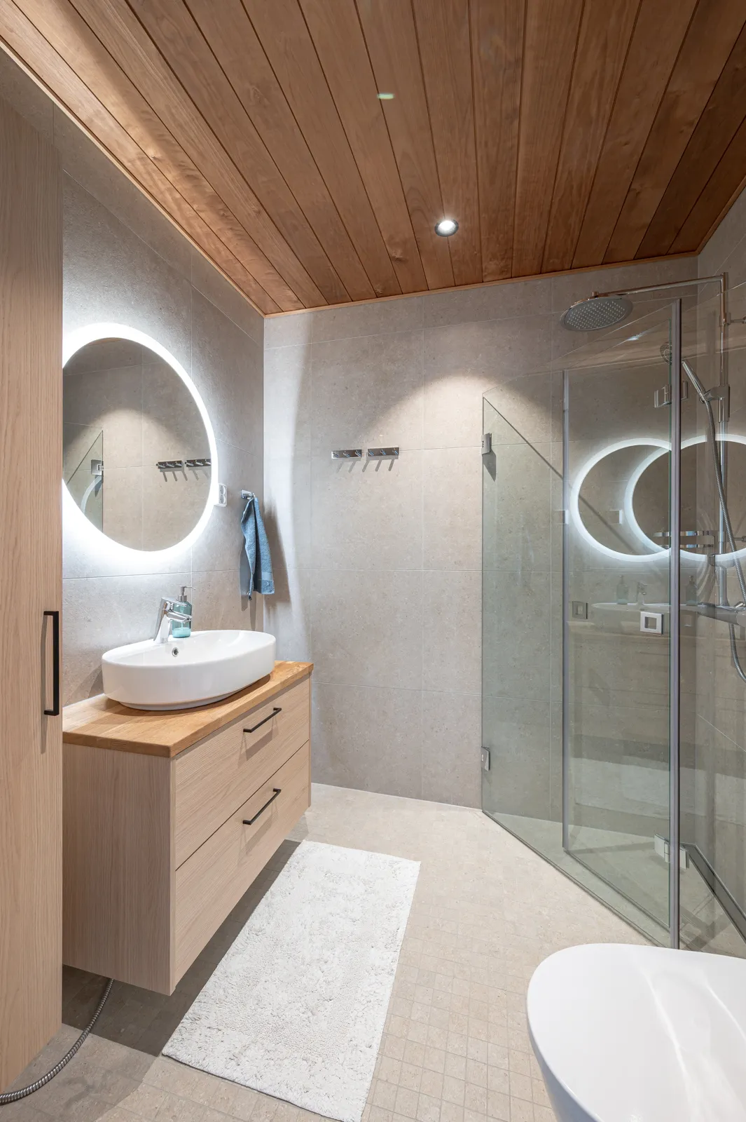 Moderni kylpyhuone, jossa on puukatto, pyöreä valaistu peili, puinen pesuallas ja lasinen suihkukaappi.