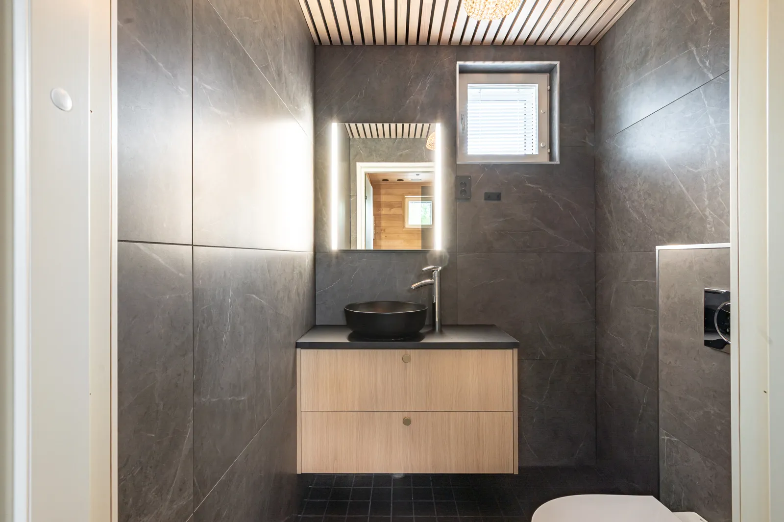 Moderni kylpyhuone, jossa on harmaat marmorilaatat, puinen katto, valaistu peili, musta pesuallas ja vaalean puun värinen allaskaappi.