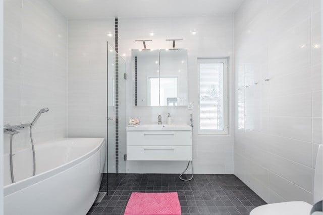 Moderni kylpyhuone valkoisilla seinälaatoilla, tummalla lattialla, valkoisella kylpyammeella ja peilikaapilla. Vaaleanpunainen matto lattialla.