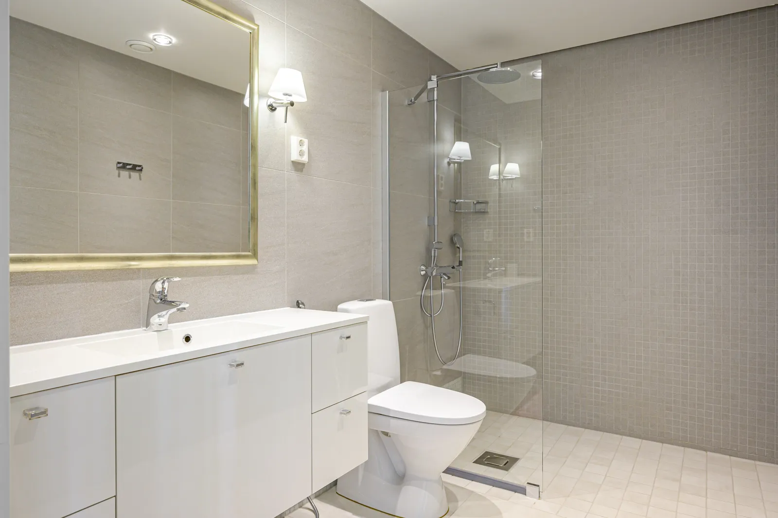 Moderni kylpyhuone valkoisella pesualtaalla, peilillä, wc:llä ja suihkulla, jossa on harmaat laatat ja kirkas valaistus.