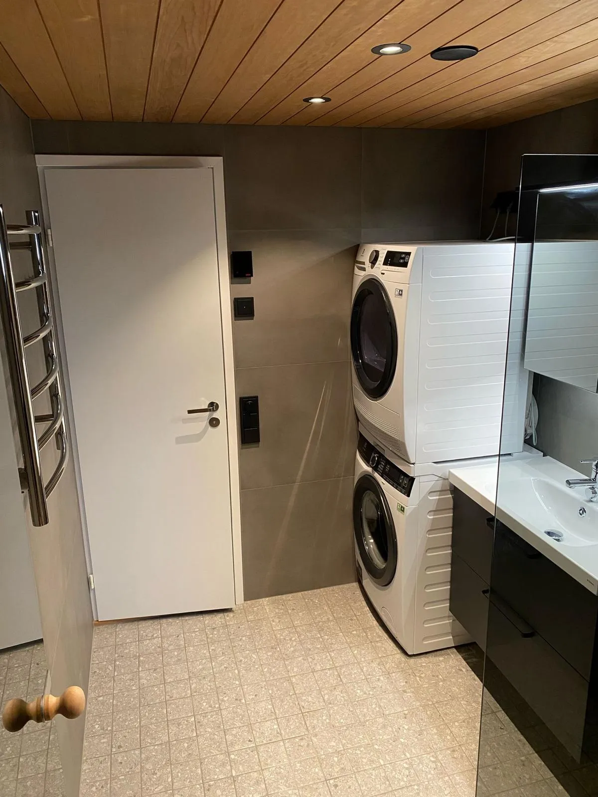 Moderni kylpyhuone, jossa on puukatto, valkoinen ovi, pyykinpesukone ja kuivausrumpu sekä harmaa laattalattia.