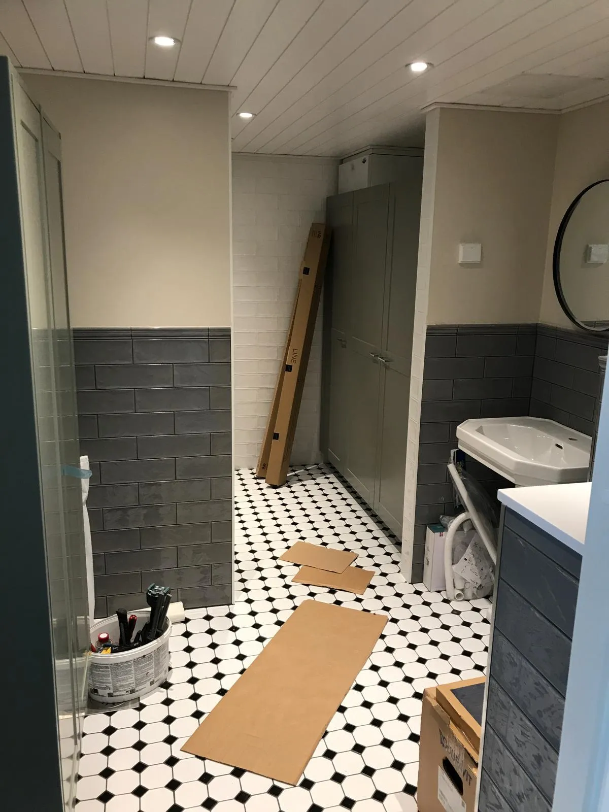 Kylpyhuoneessa on mustavalkoinen lattia, harmaat seinälaatat ja pahvilaatikoita lattialla. Valoisa tila, jossa on katossa upotetut valaisimet.