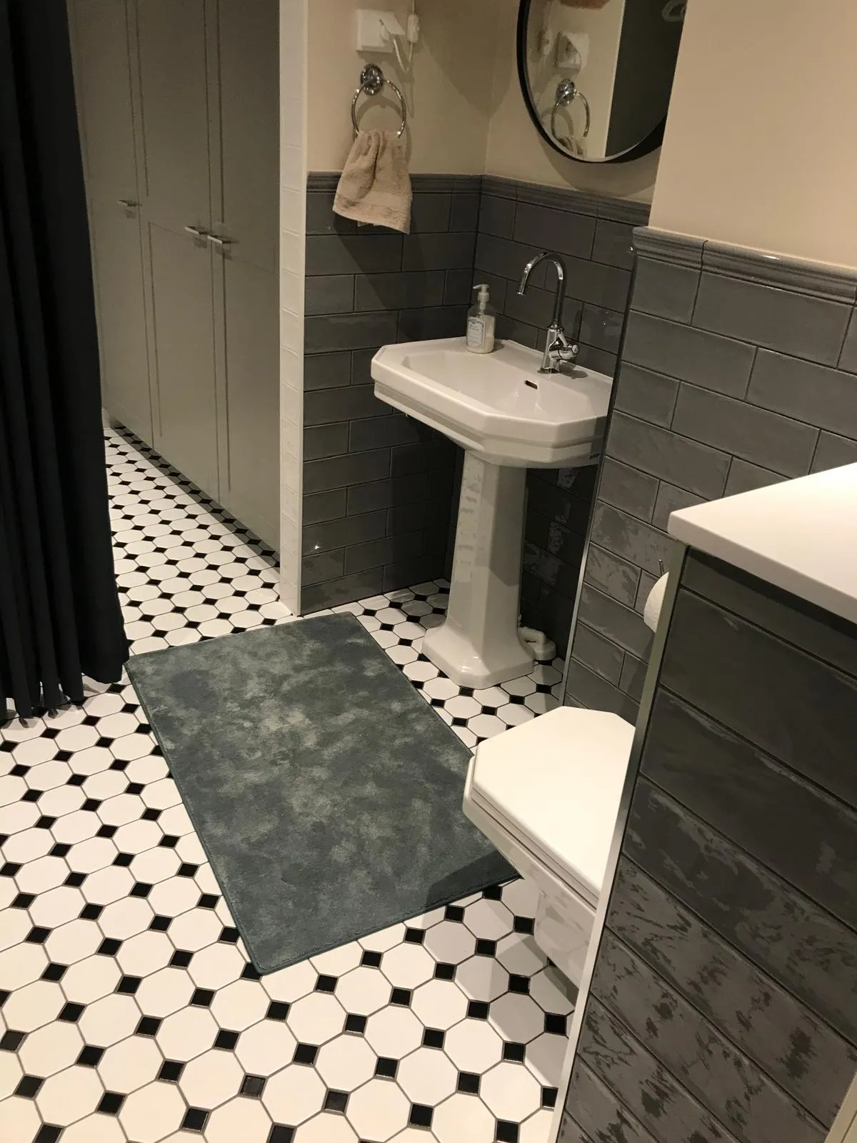 Kylpyhuoneen sisustus, jossa on mustavalkoinen laattalattia, harmaa matto, valkoinen pesuallas ja wc, sekä harmaat seinälaatat.