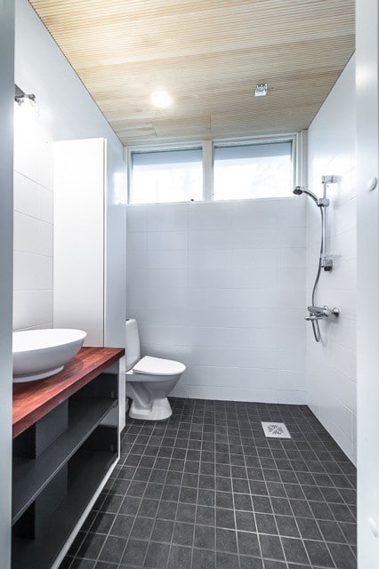 Moderni kylpyhuone, jossa on puukatto, valkoiset seinät, tummat lattialaatat, valkoinen pesuallas puutasolla ja wc-istuin.