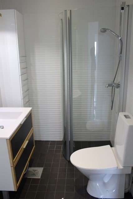 Kylpyhuoneen sisustus, jossa on valkoinen pesuallas, peilikaappi, suihkukaappi ja valkoinen WC-istuin tummalla laattalattialla.