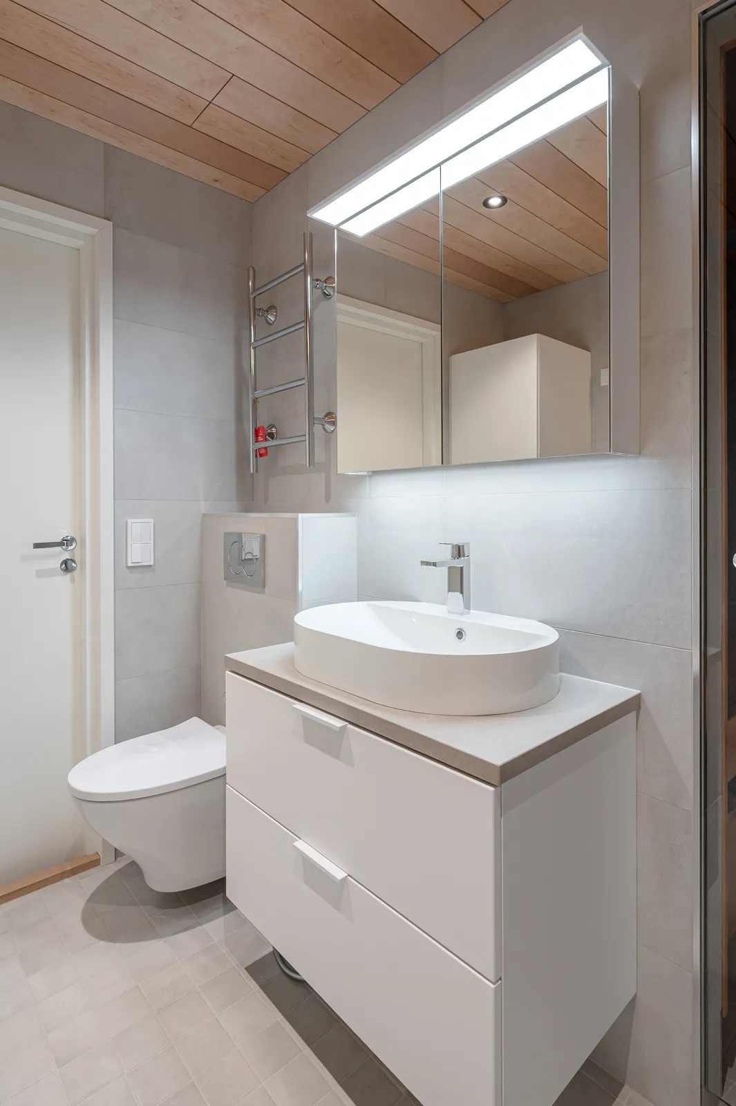 Moderni kylpyhuone, jossa on valkoinen pesuallas, peilikaappi valolla ja puinen katto.