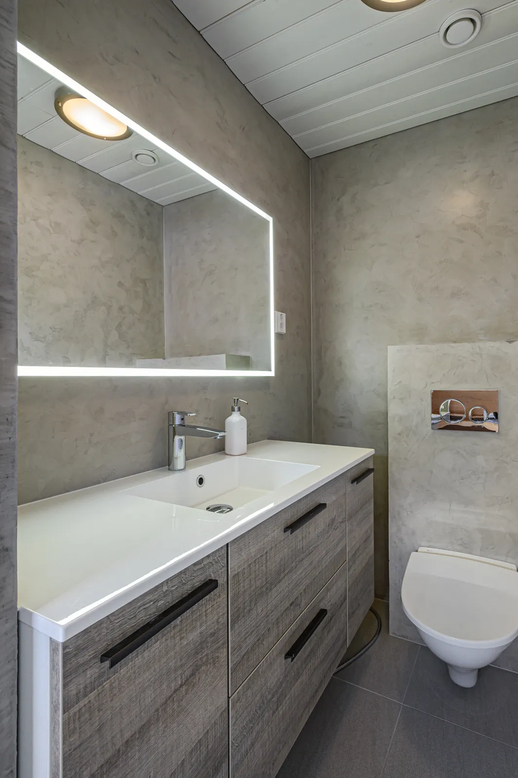 Kylpyhuoneen sisustus, jossa on harmaat seinät, valkoinen katto, valaistu peili, valkoinen pesuallas, puinen allaskaappi ja seinä-wc.