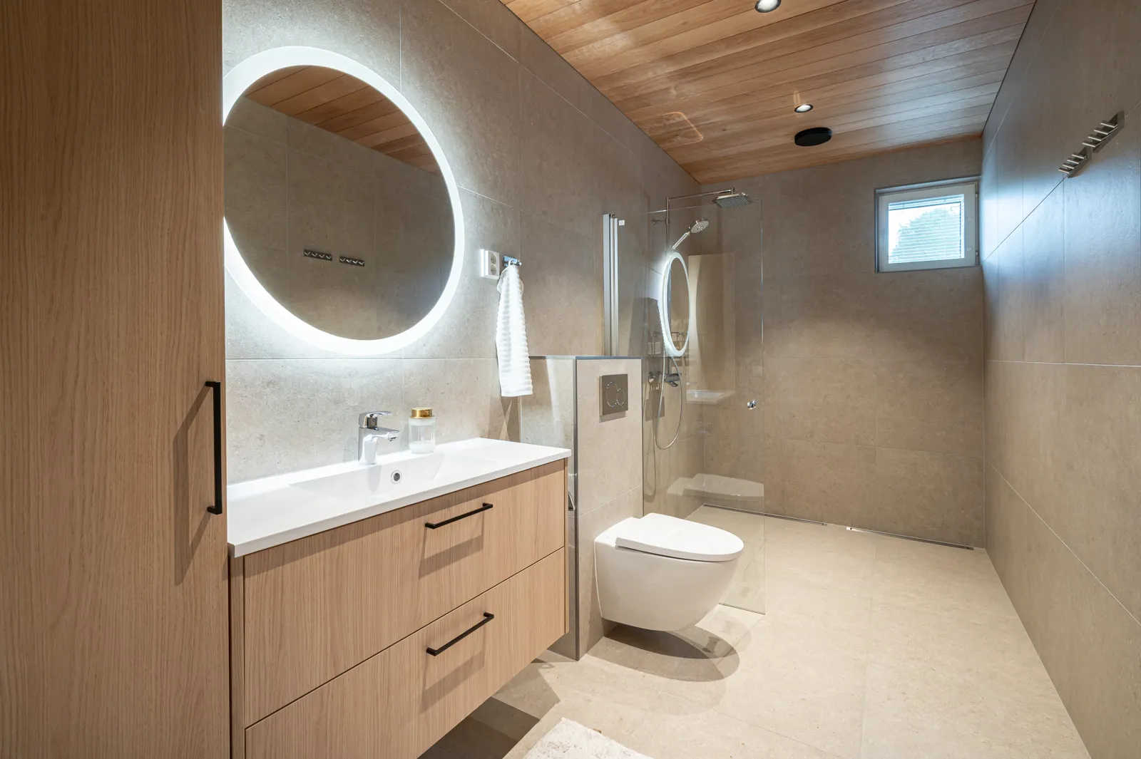 Moderni kylpyhuone, jossa on puukatto, pyöreä peili, puinen pesuallas ja seinään asennettu wc. Valoisa ja tyylikäs sisustus.