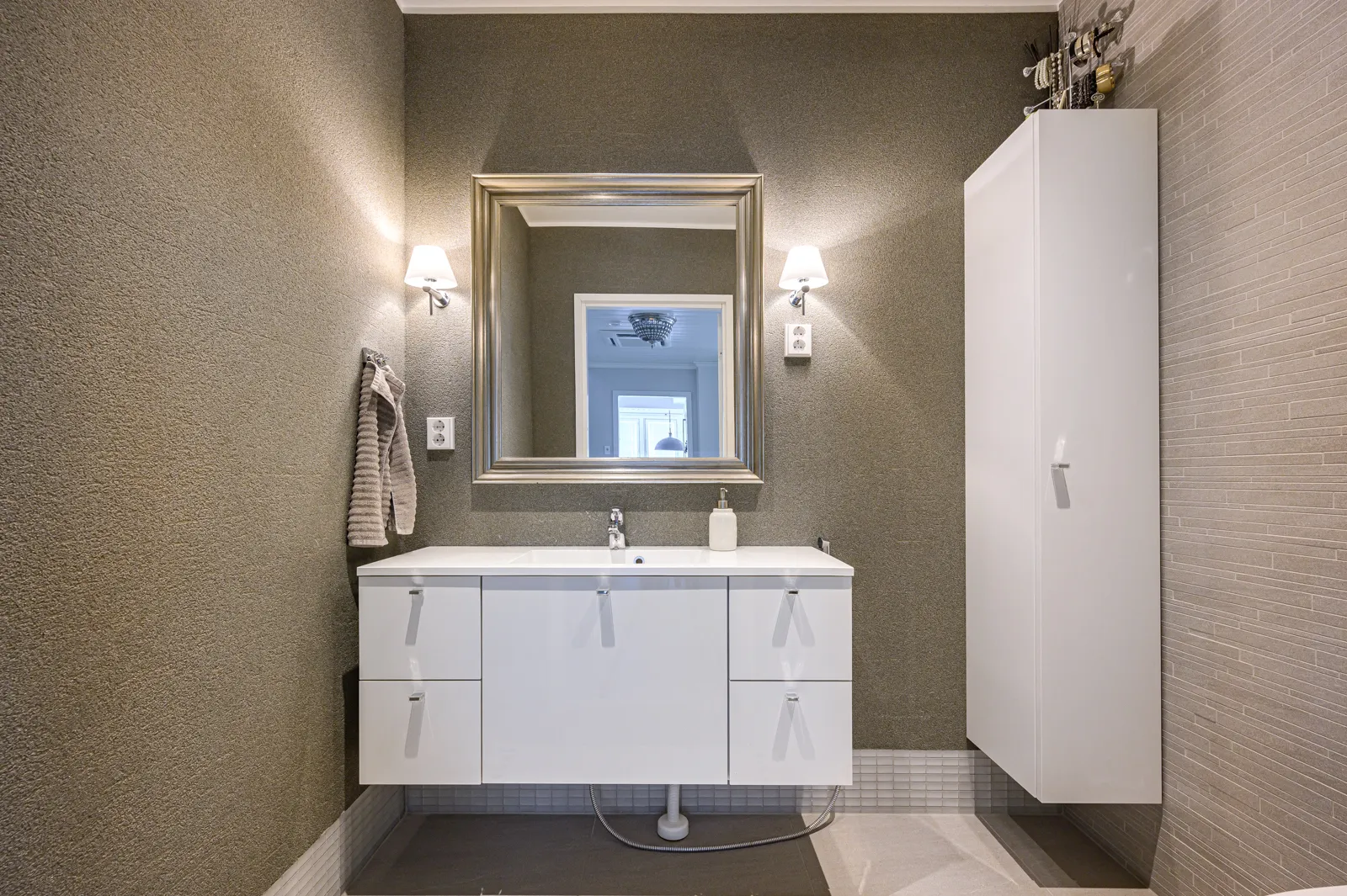 Kylpyhuoneen sisustus, jossa on valkoinen pesuallas, peili, seinävalaisimet ja valkoinen kaappi. Seinät ovat harmaat ja valaistus on lämmin.