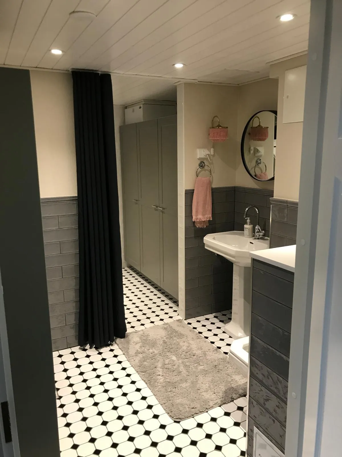 Kylpyhuoneen näkymä, jossa on harmaa kaappi, musta verho, valkoinen pesuallas ja mustavalkoinen lattia.