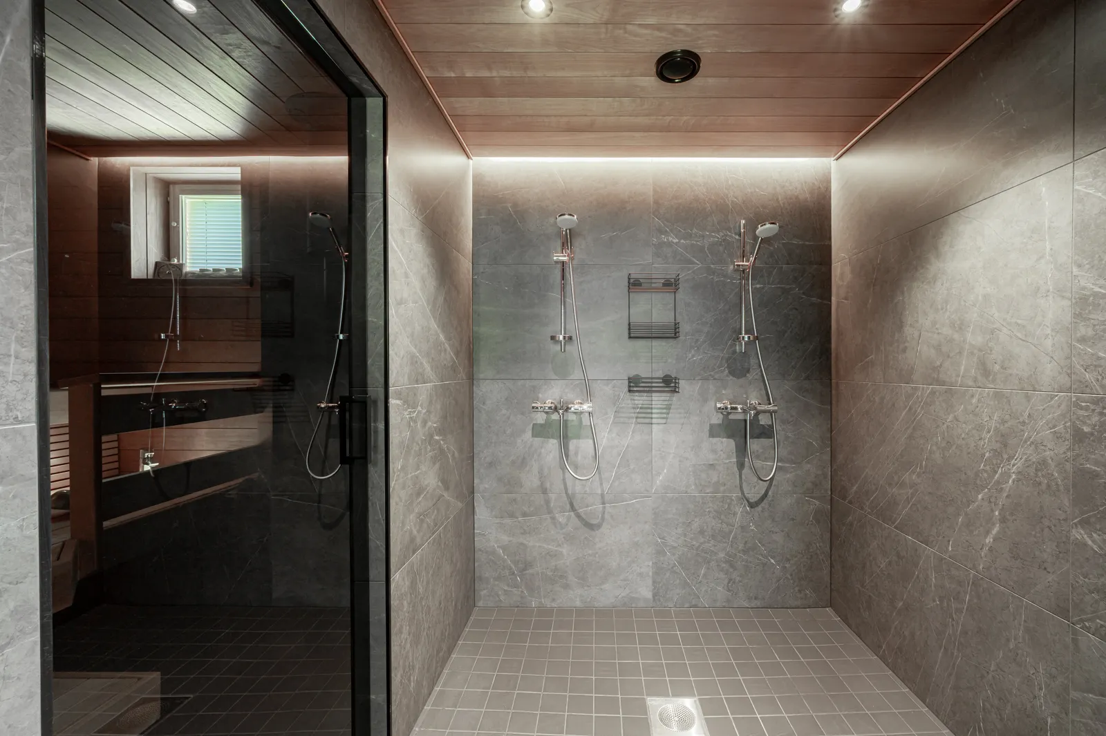 Moderni kylpyhuone, jossa on harmaat marmorilaatat, puinen katto ja kaksi suihkua. Saunan lasiovi näkyy vasemmalla.