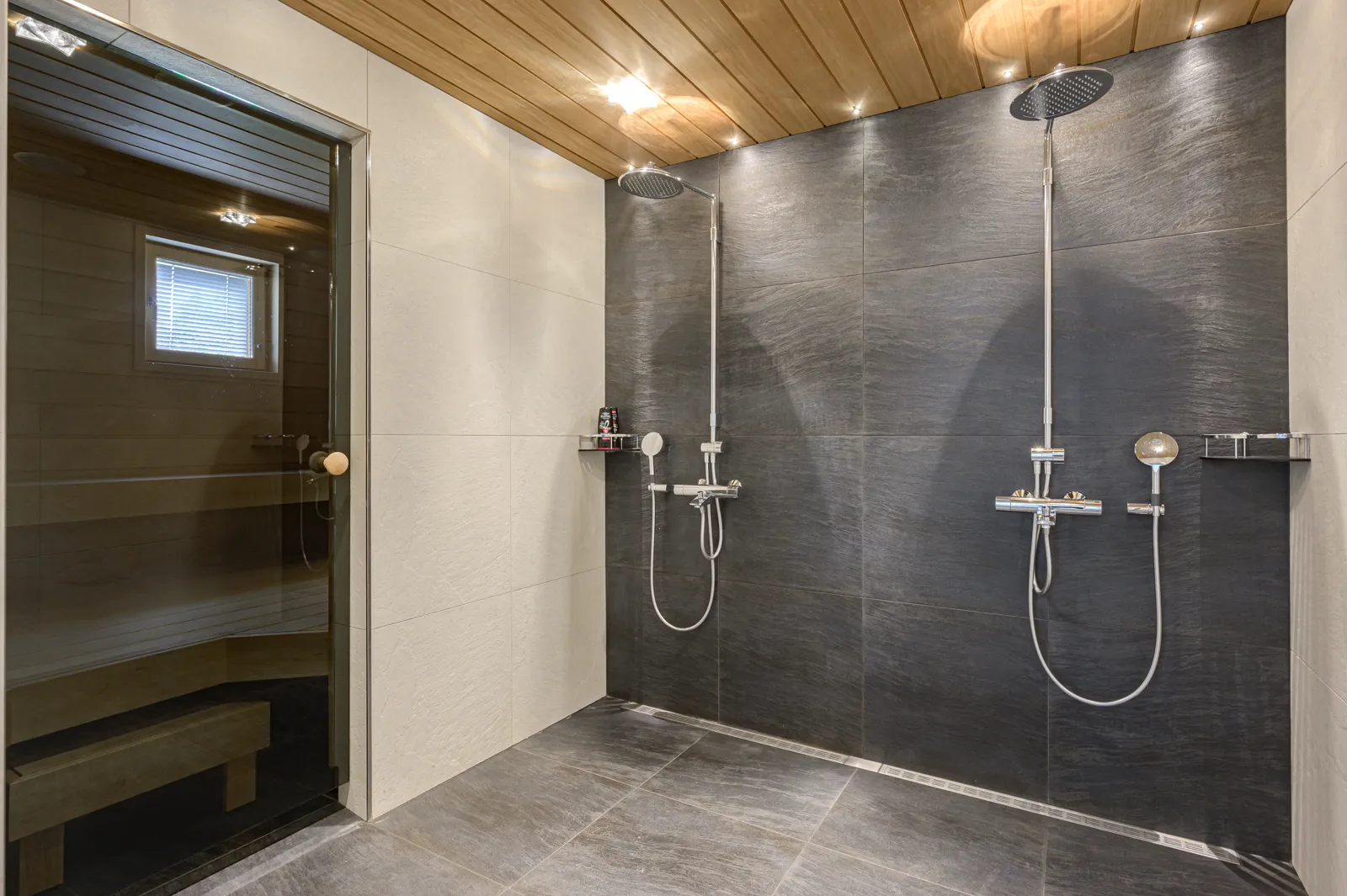Moderni kylpyhuone, jossa on kaksi suihkua, sauna lasiovella, puinen katto ja harmaat laatat.