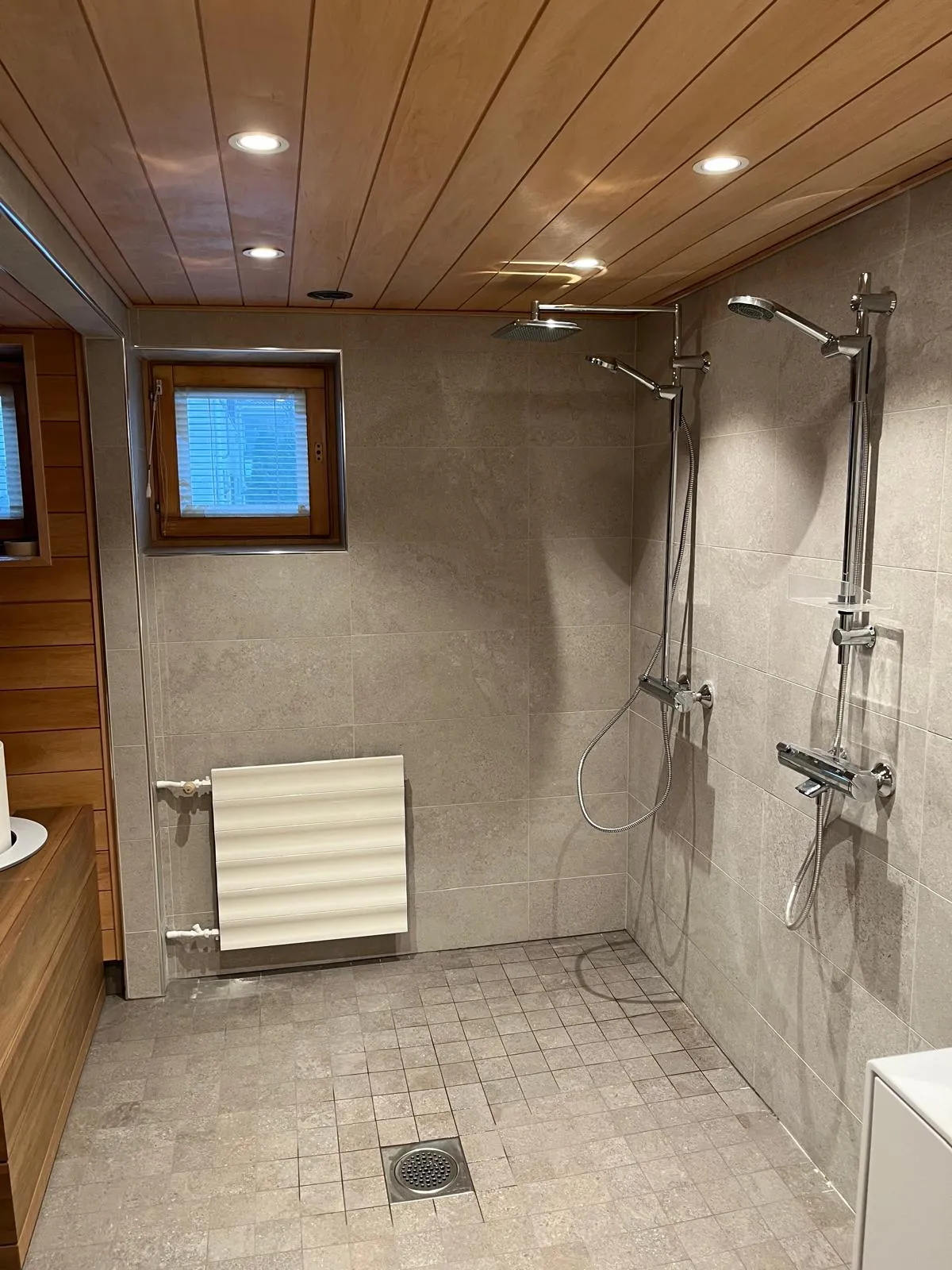 Kylpyhuoneen sisustus, jossa on puukatto, harmaat laatat, kaksi suihkua ja valoisa ikkuna.