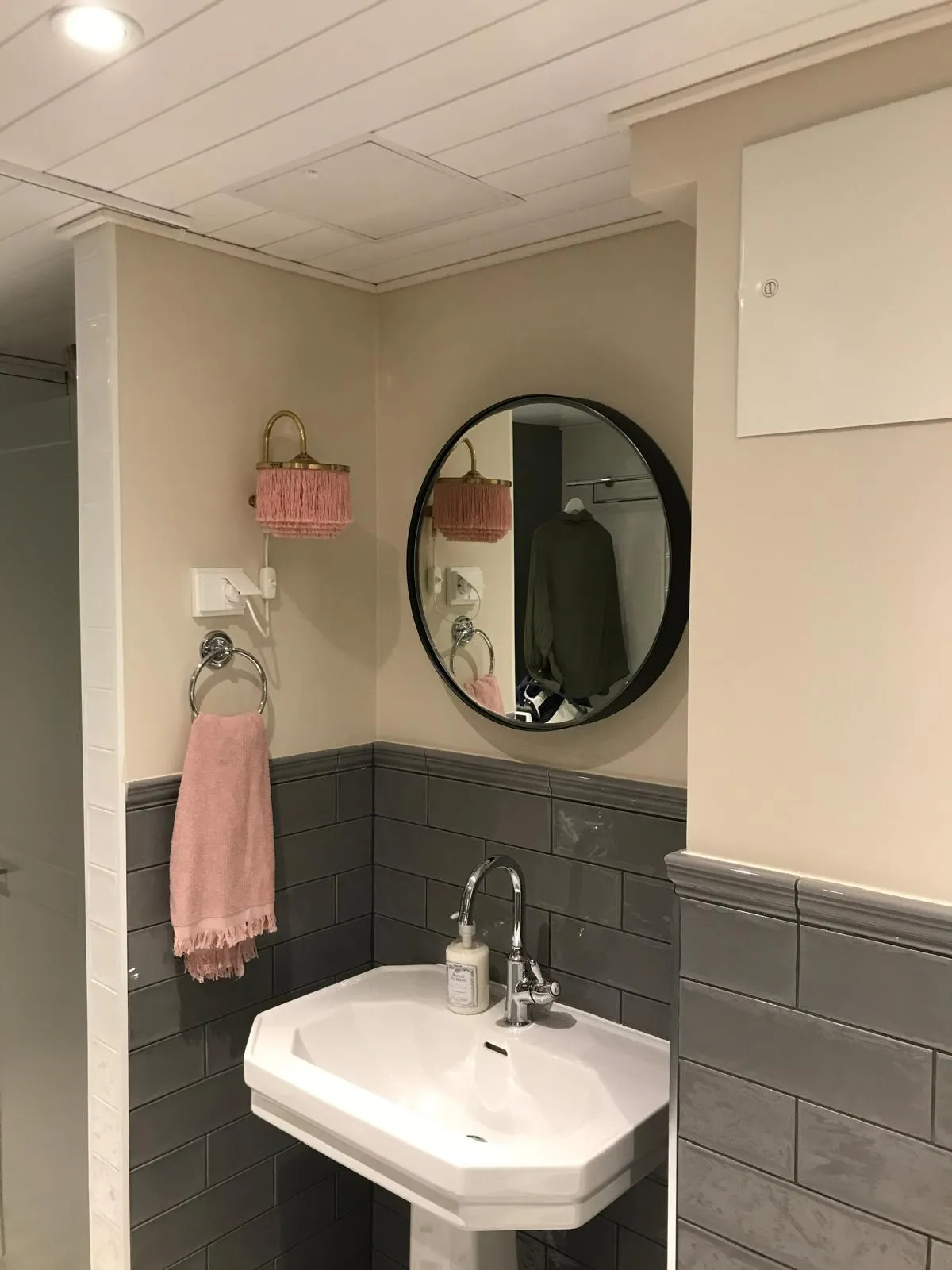 Kylpyhuoneessa on valkoinen pesuallas, harmaat laatat, pyöreä peili ja vaaleanpunainen valaisin. Seinällä on valkoinen sähkökaappi ja katossa valkoinen paneeli.