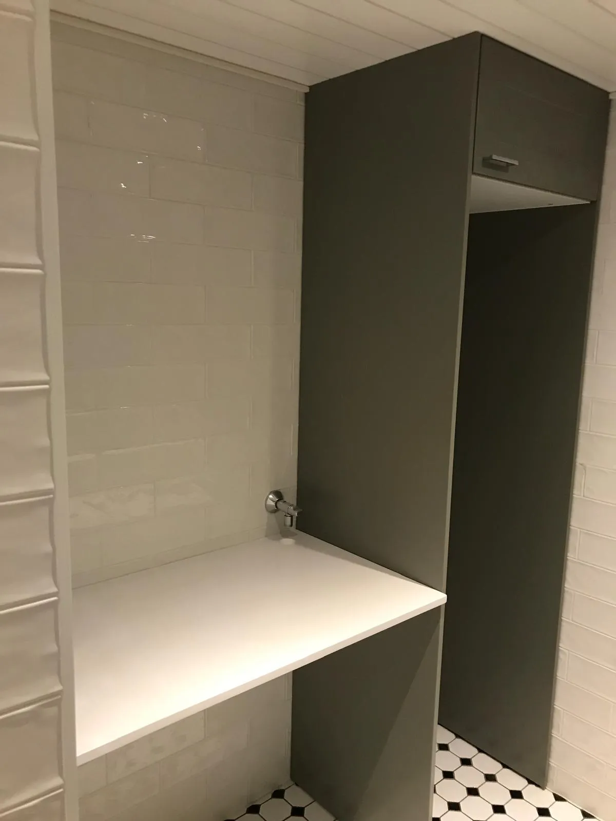 Kylpyhuoneen sisustus, jossa on valkoinen taso, harmaa kaappi ja mustavalkoinen lattia. Seinät on laatoitettu valkoisilla suorakaiteen muotoisilla laatoilla.