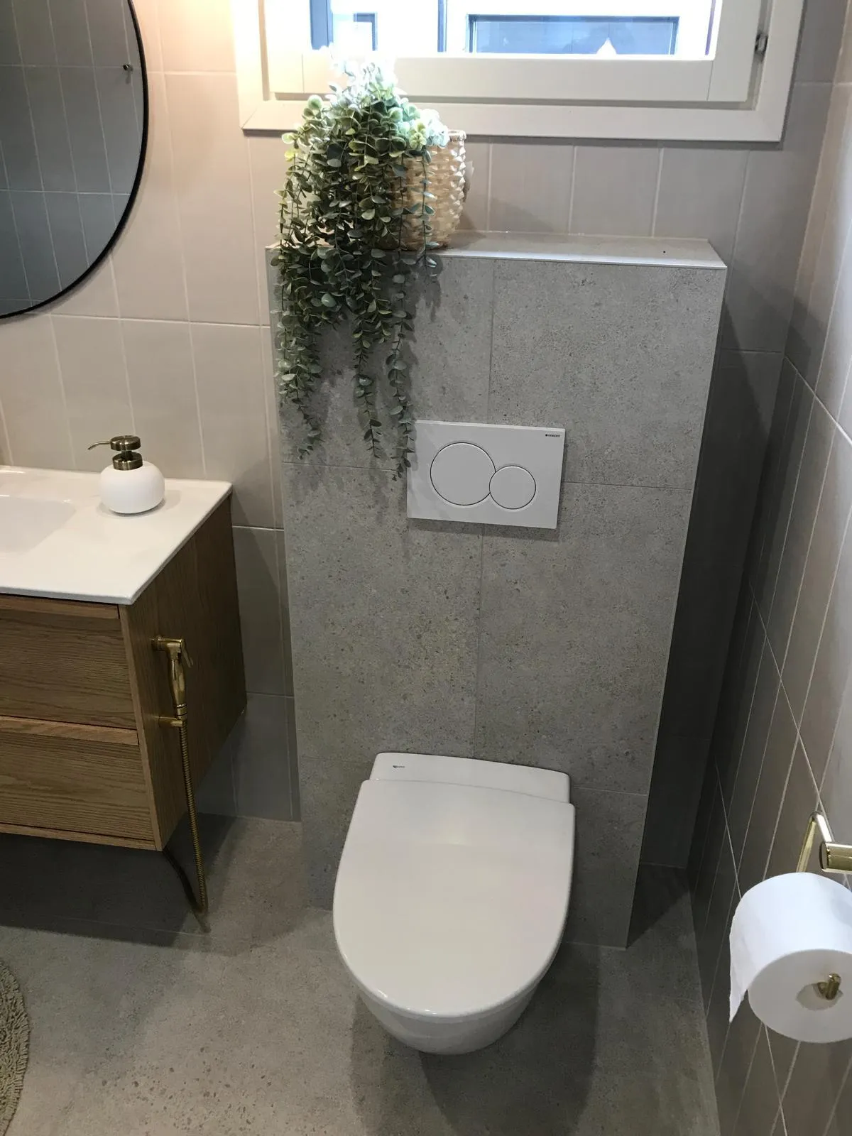 Moderni kylpyhuone, jossa on valkoinen wc, harmaat laatat, puinen pesuallas, viherkasvi ja pyöreä peili.