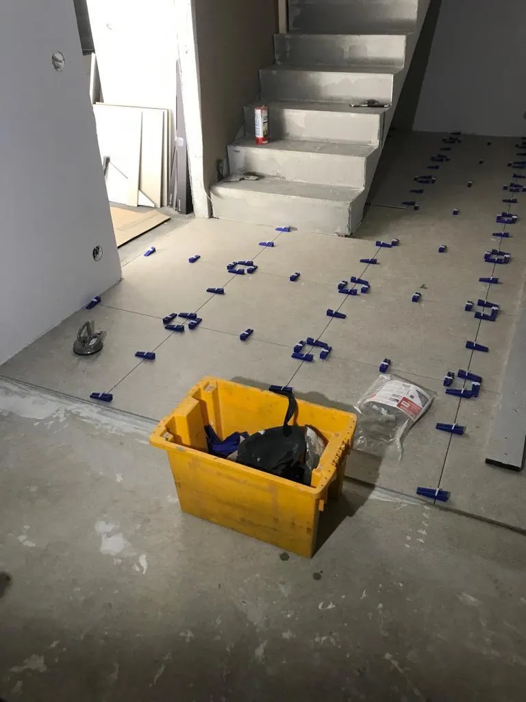 Keltaisessa laatikossa on työkaluja, ja lattialla on sinisiä laattojen asennusvälineitä portaiden alla, valaistus on himmeä.