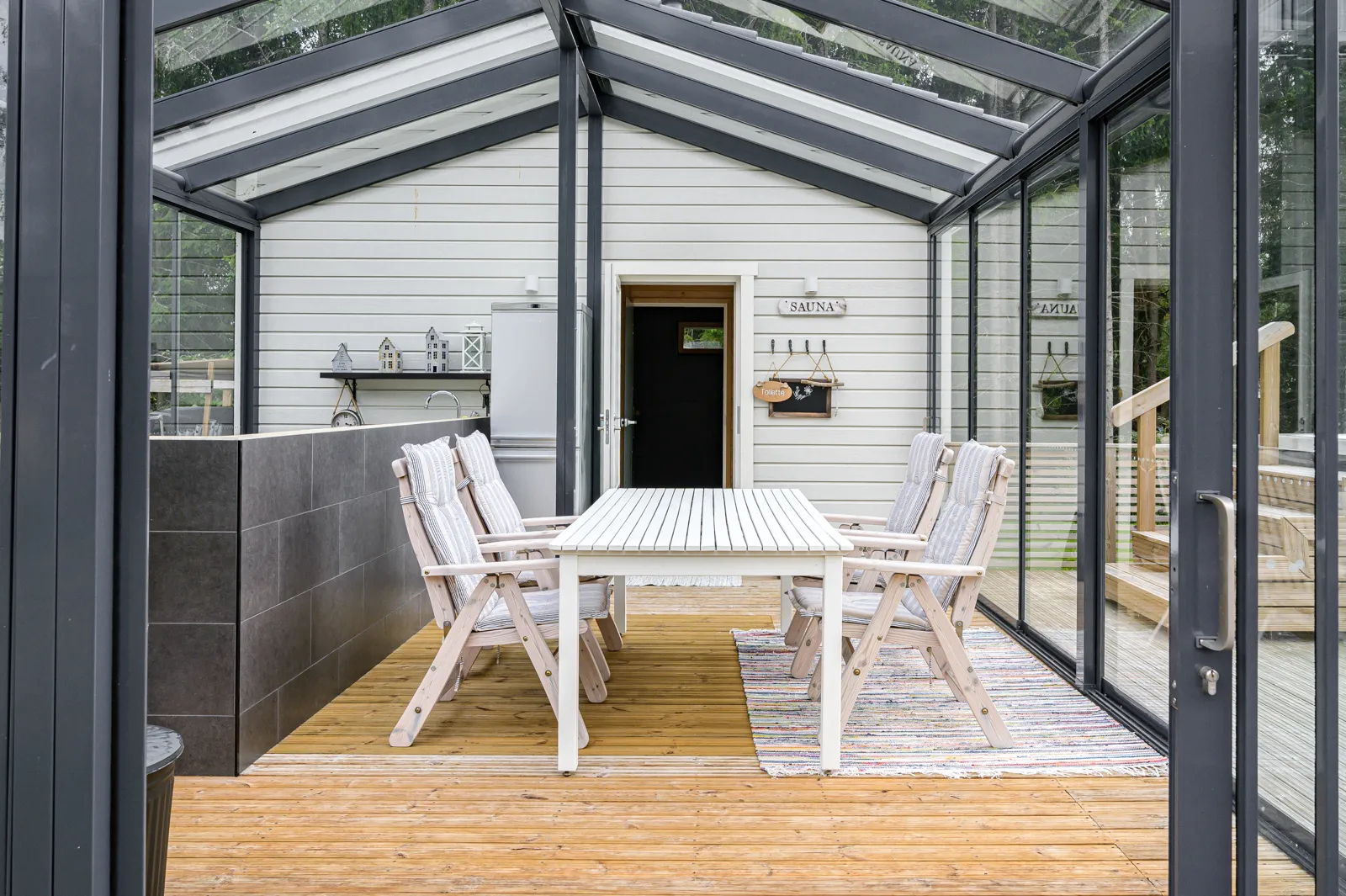 Valoisassa lasikuistissa on puinen ruokapöytä tuoleineen maton päällä, sekä saunaan johtava ovi ja pieni keittiönurkkaus.