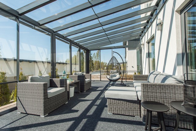 Valoisassa lasitetussa terassissa on rottinkiset istuinryhmät, riipputuoli ja tumma matto, auringonvalo siivilöityy lasikaton läpi.