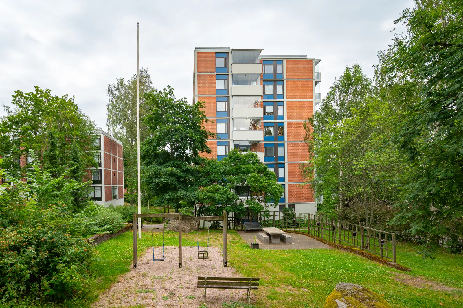 Ulkona on leikkipaikka, jossa on keinut, penkki ja pöytäryhmä, taustalla kerrostaloja pilvisen sään alla.