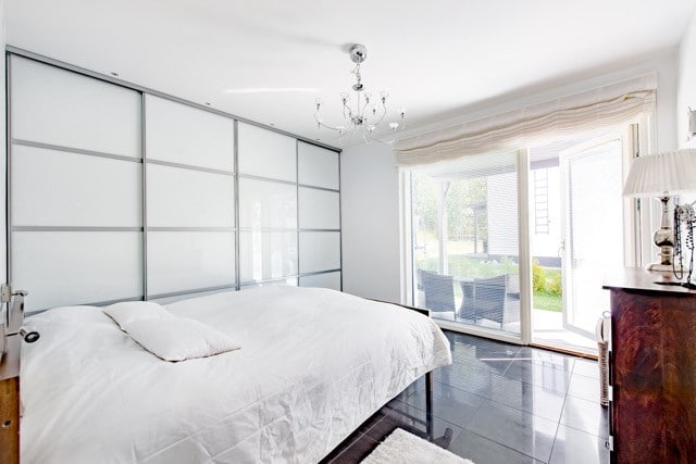 Vaaleassa makuuhuoneessa on suuri sänky valkoisilla liinavaatteilla, liukuovet, kattokruunu ja avoin parvekeovi, josta näkyy ulos.