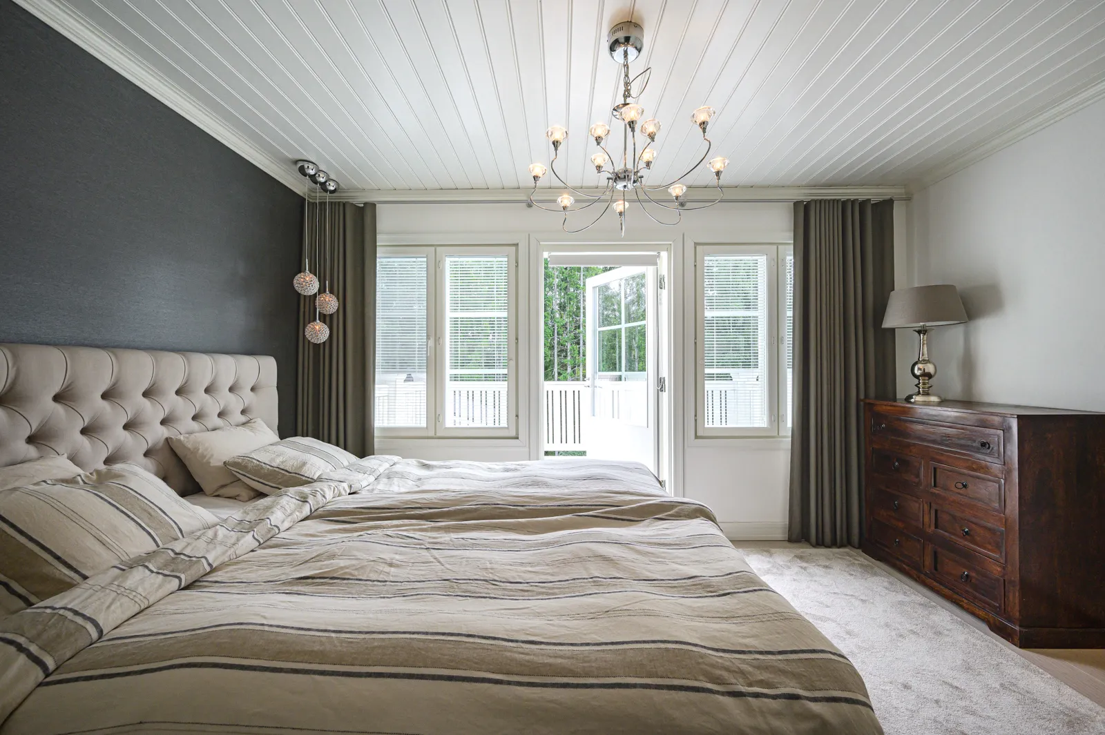 Makuuhuoneessa on parivuode, yöpöytä ja avoin parvekkeen ovi, valaistuksena kattokruunu ja yöpöydän lamppu.