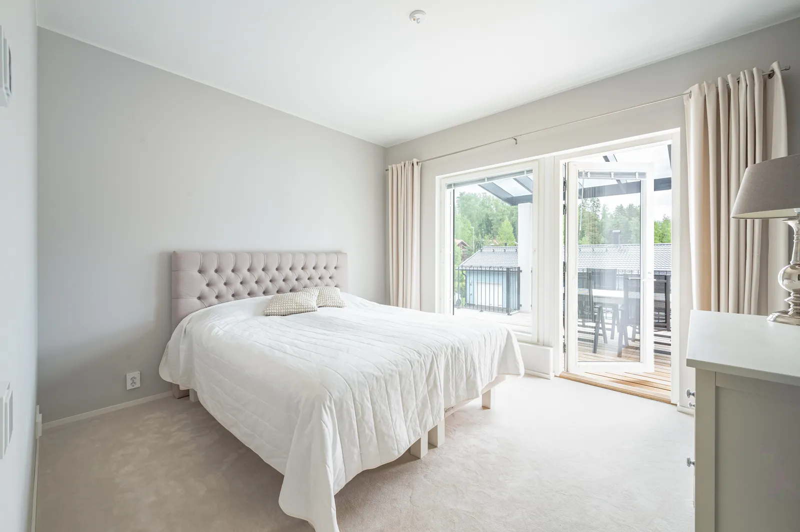 Vaaleassa makuuhuoneessa on sängynpääty, valkoinen päiväpeite, ikkunat ja parveke. Huoneessa on myös yöpöytä ja lampunvarjostin.