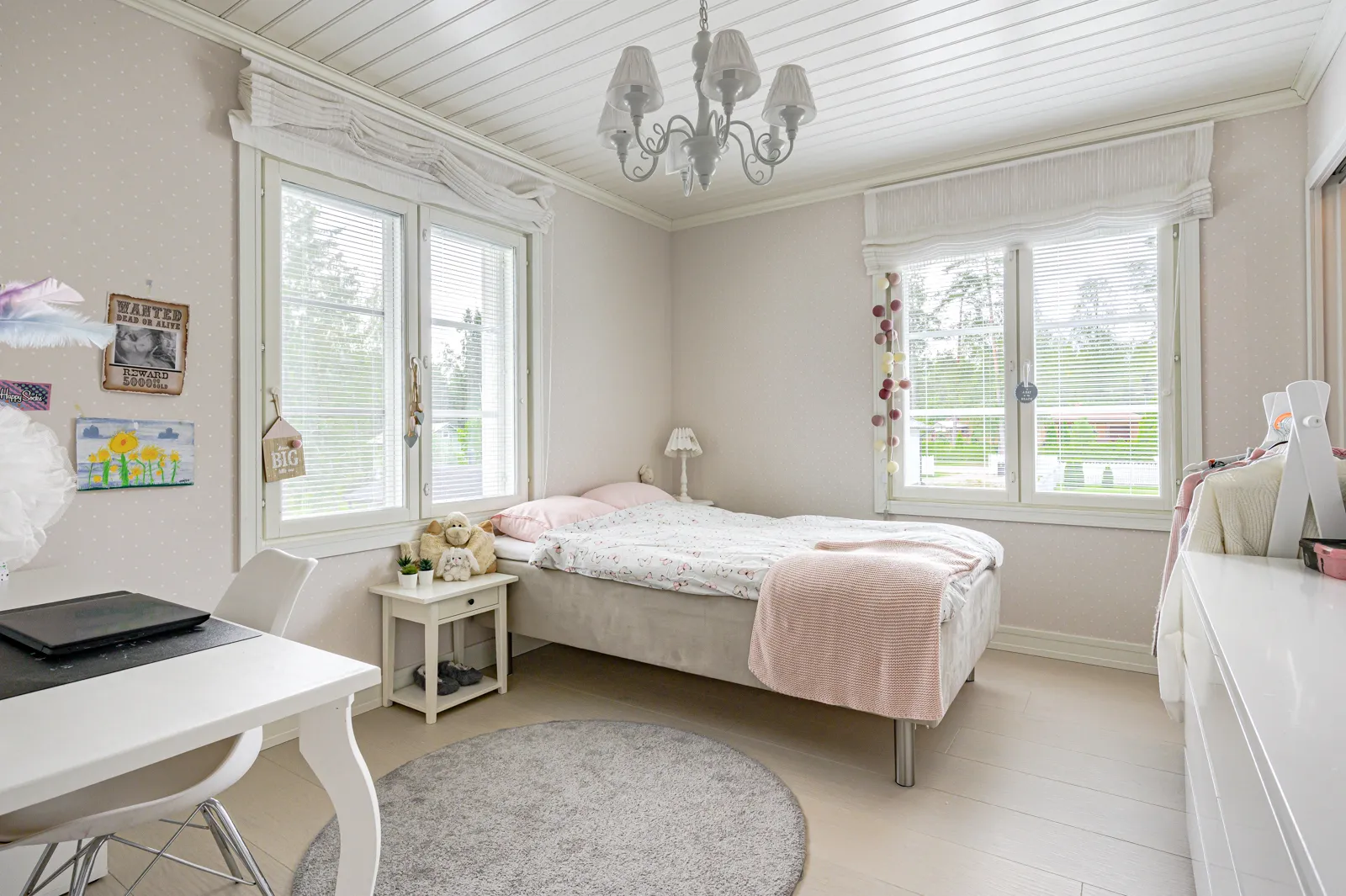 Vaaleassa makuuhuoneessa on sänky, työpöytä ja pyöreä matto. Huoneessa on kaksi ikkunaa, joissa on valkoiset sälekaihtimet. Katossa roikkuu valkoinen kattokruunu. Seinillä on tauluja ja koristeita.