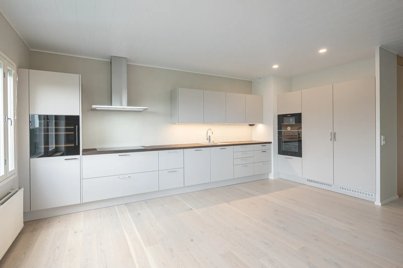 Vaalea, moderni keittiö, jossa on valkoiset kaapit, puinen työtaso ja ruostumattomasta teräksestä valmistettu liesituuletin. Lattiat ovat vaaleaa puuta ja valaistus on kirkas.