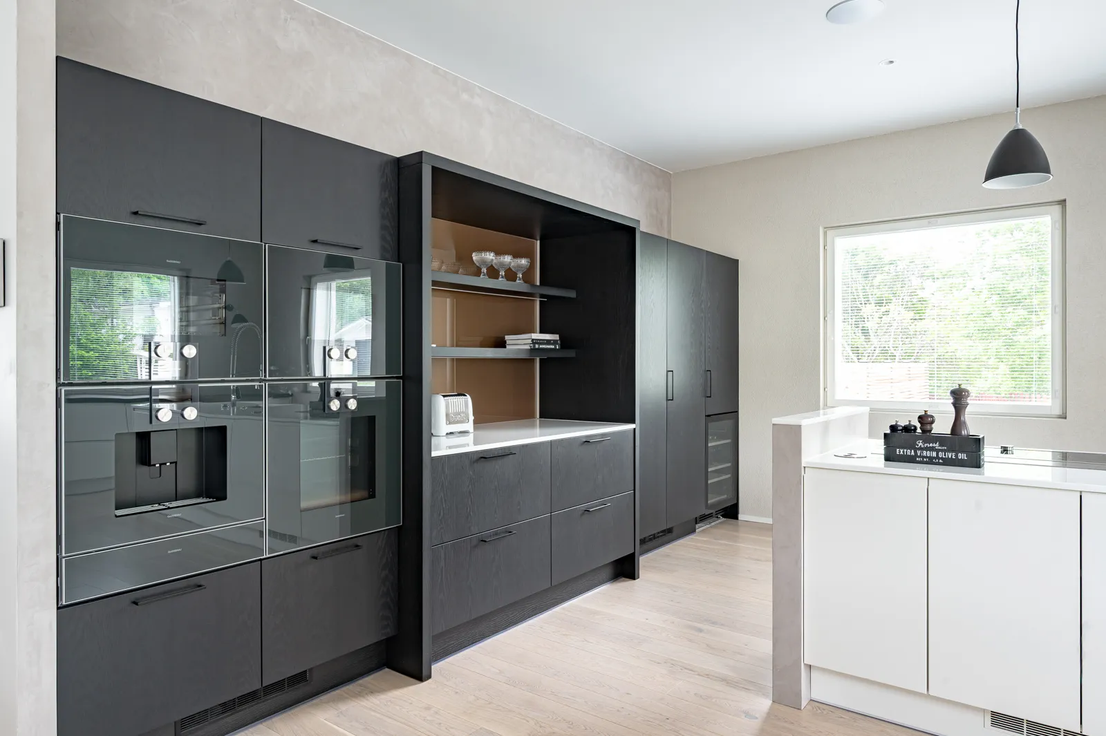 Moderni keittiö, jossa on tummat puukaapit, integroidut kodinkoneet, valkoinen työtaso ja vaaleat puulattiat. Ikkunasta tulee luonnonvaloa.
