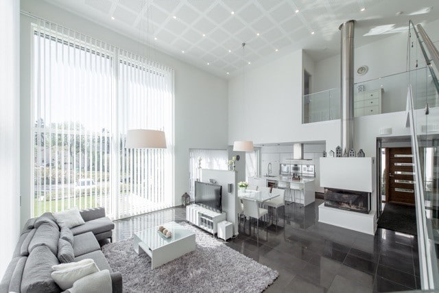 Moderni olohuone, jossa harmaa sohva, valkoinen sohvapöytä, suuret ikkunat ja avokeittiö, valaistus on kirkas ja tila avara.