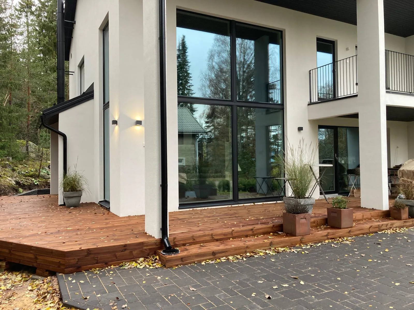 Moderni, valkoinen talo, jossa on suuret ikkunat, puinen terassi ja syksyn lehtiä maassa. Ulkovalot valaisevat seinää.