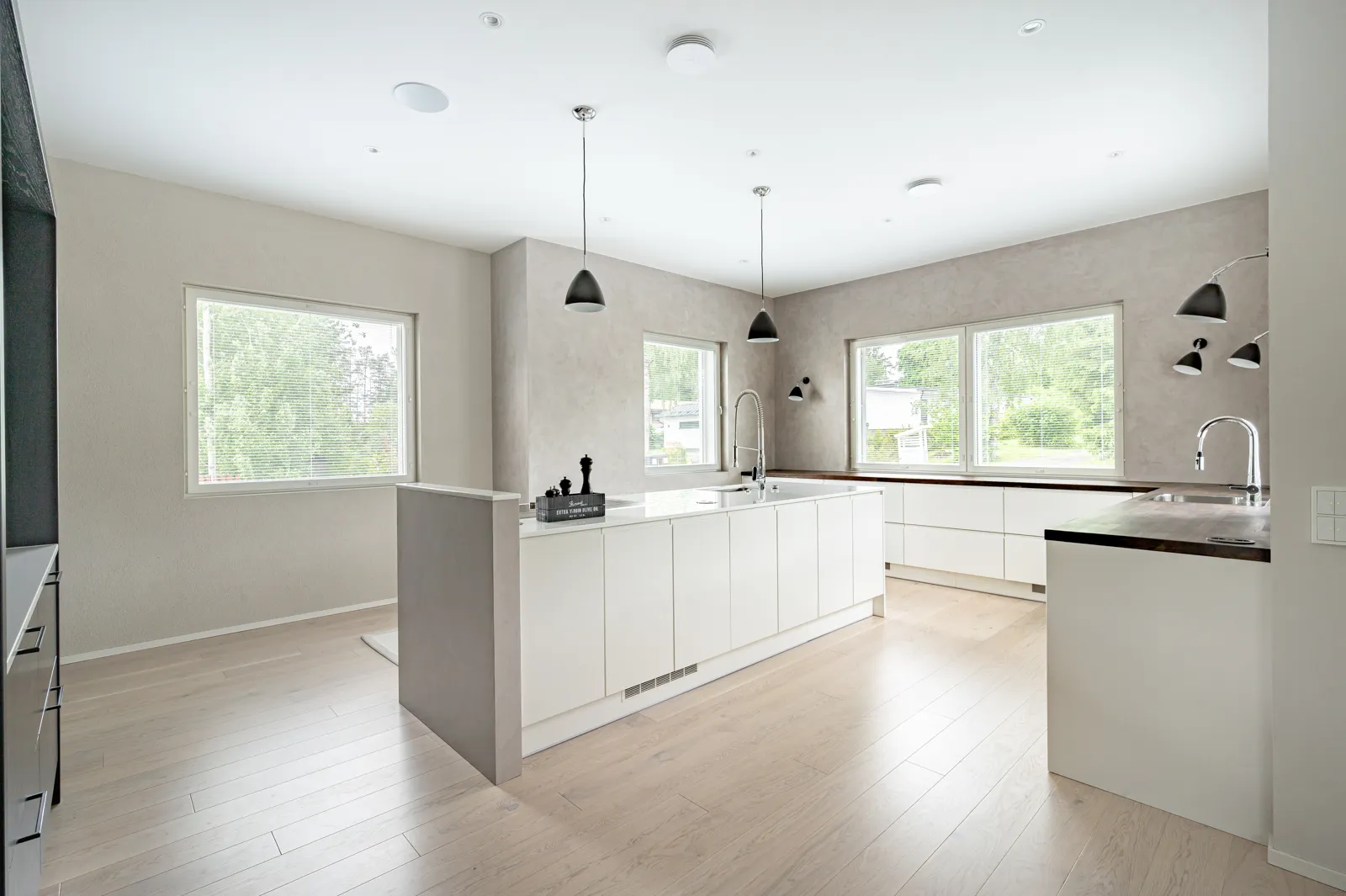 Moderni keittiö, jossa on valkoiset kaapit, vaaleat puulattiat ja suuret ikkunat, jotka tuovat luonnonvaloa. Mustat riippuvalaisimet ja seinävalaisimet lisäävät tyylikkään kosketuksen.