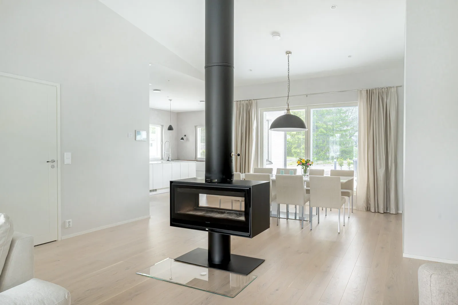 Moderni olohuone, jossa on musta takka, vaaleat puulattiat ja valkoinen ruokailutila ikkunoiden äärellä.