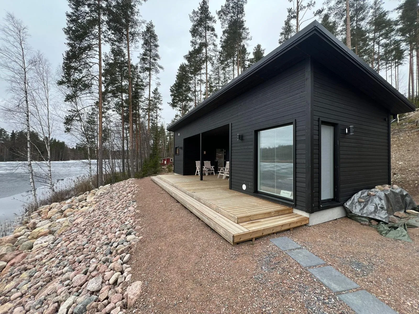Moderni musta mökki puuterassilla järven rannalla, kivikkoinen ranta, harmaa taivas.