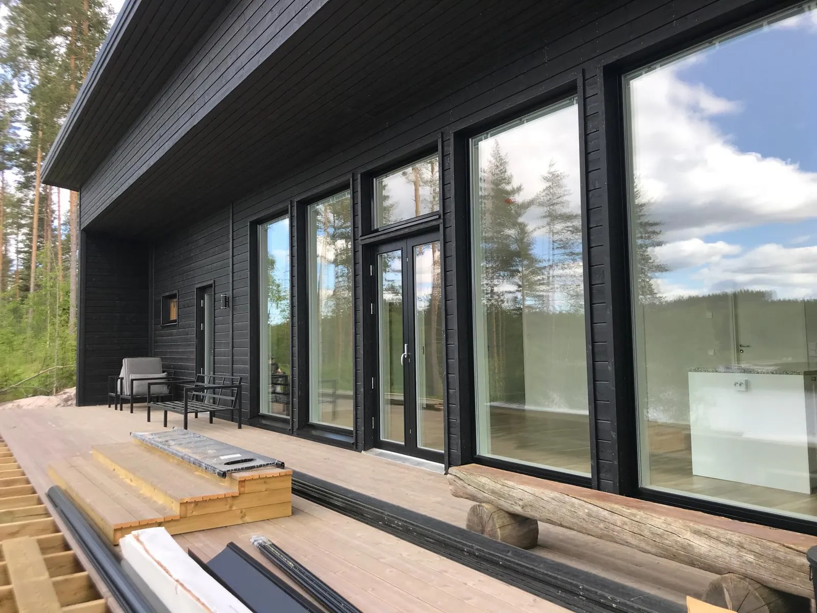 Moderni musta puutalo, jossa suuret ikkunat heijastavat maisemaa, puinen terassi ja ulkokalusteet valoisassa säässä.