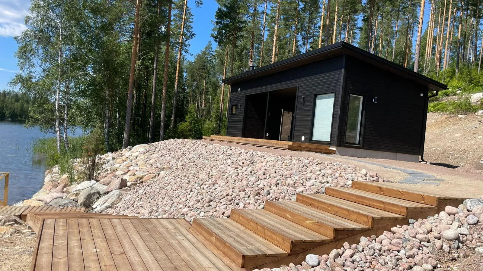 Moderni, musta puutalo kivikkoisella rinteellä järven rannalla, puinen terassi ja portaat etualalla, aurinkoinen sää.