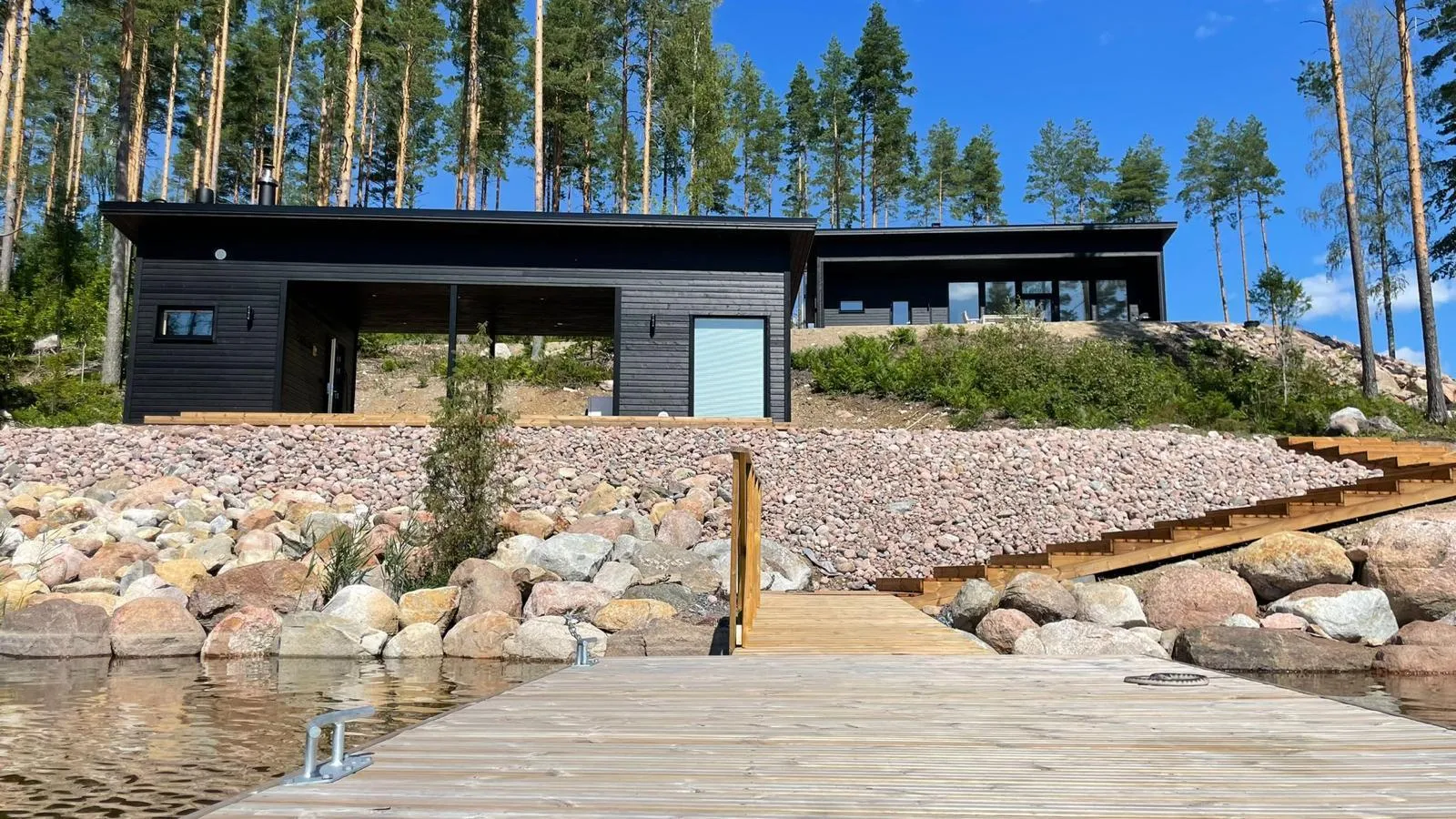 Moderni musta talo kivikkoisella rannalla, puinen laituri etualalla, taustalla korkeita puita ja sininen taivas.