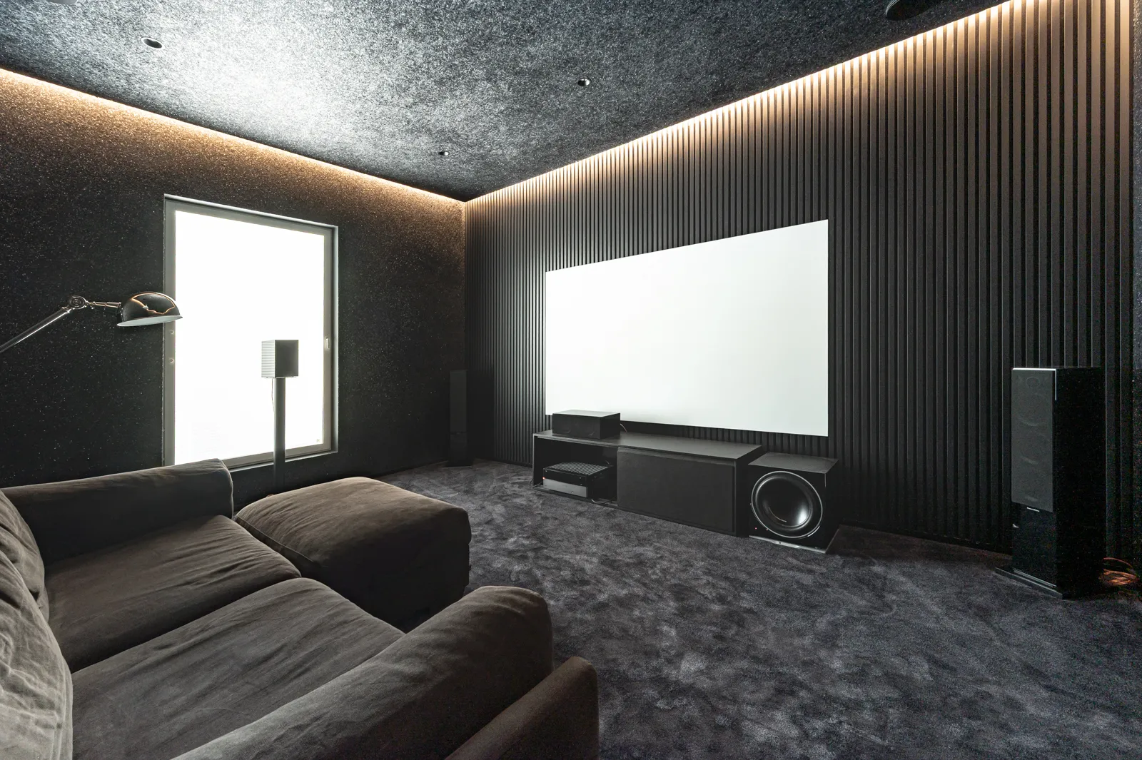 Musta kotiteatterihuone, jossa on tumma sohva, valkoinen valkokangas ja epäsuora valaistus.